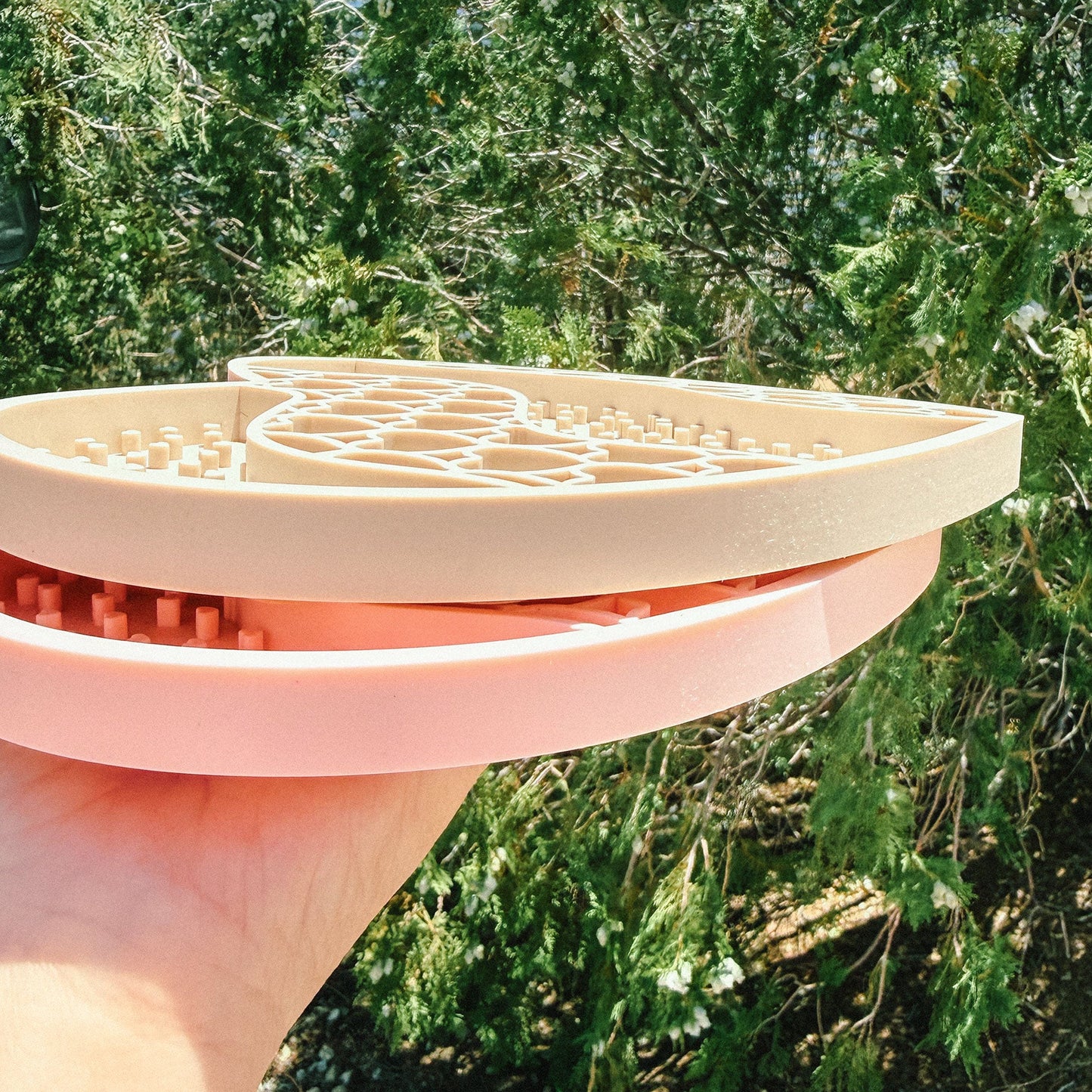 A hand holding a light pink silicone kitchen tool, likely a cookie lift or pastry helper, with a white plastic insert, set against a background of green outdoor foliage.