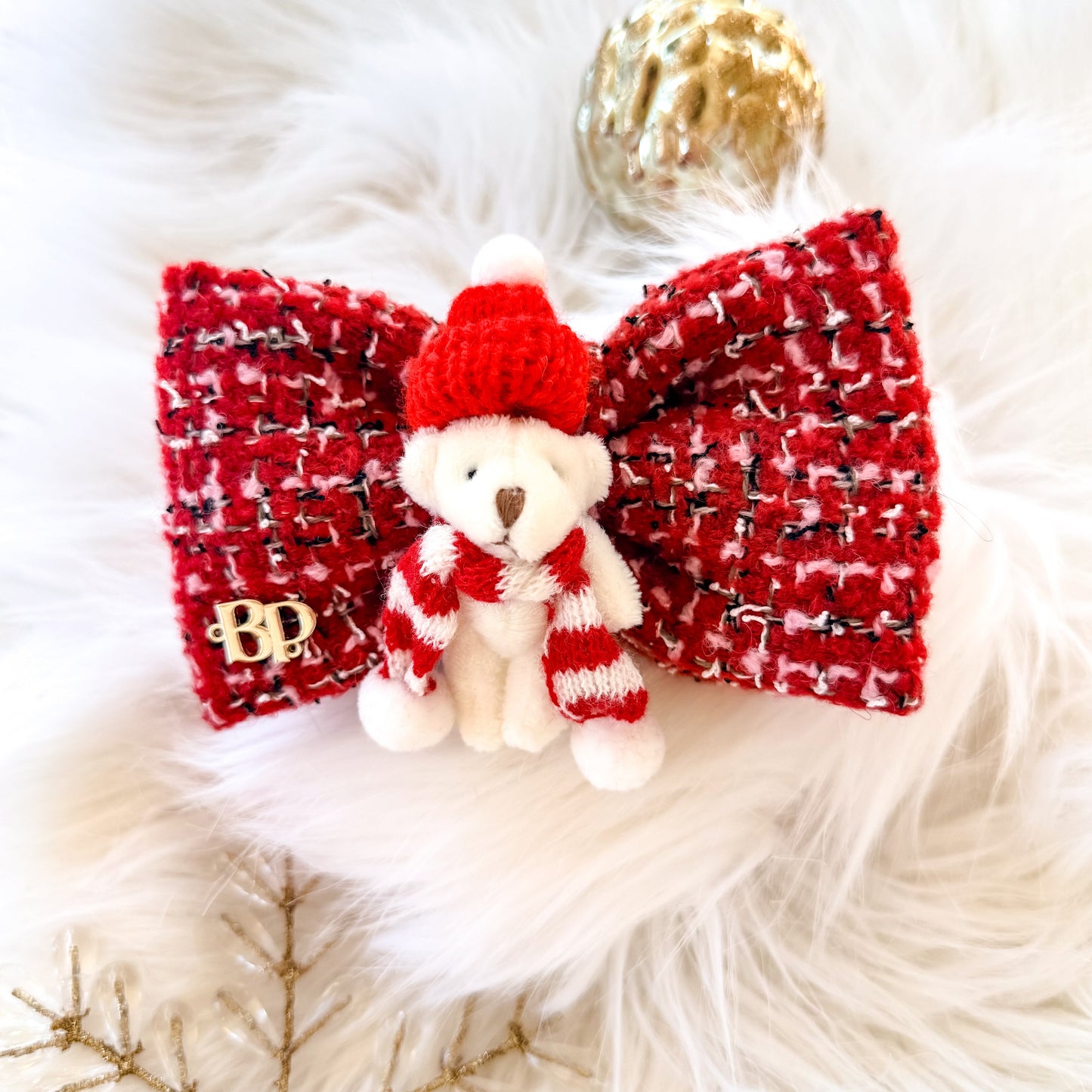 A festive red and white tweed pet bow tie featuring a small white mini plush teddy bear wearing a red knit hat and striped scarf, displayed on a white faux fur surface with holiday snowflake crystal and gold ornaments