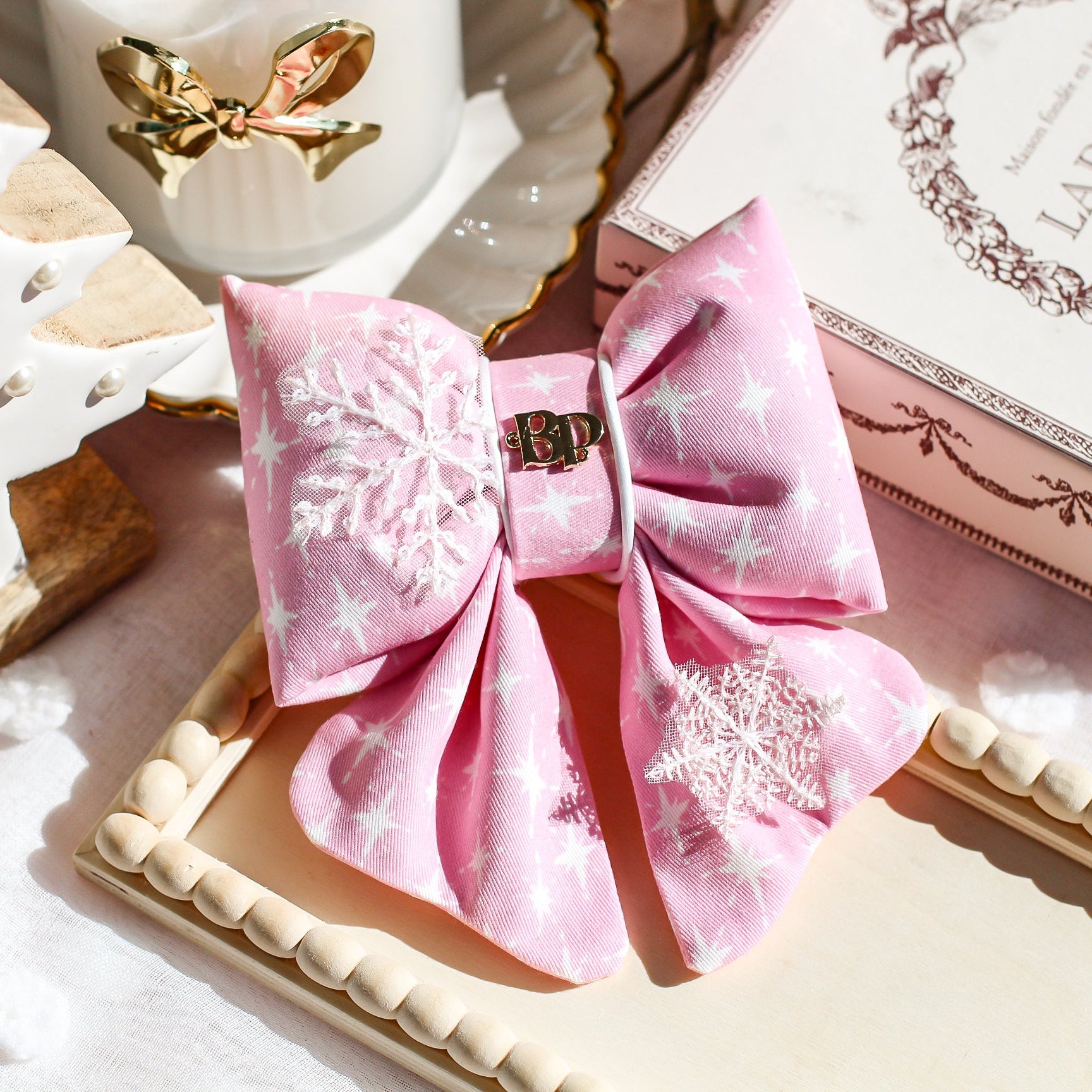 Decorative pink bow with snowflake patterns on a cookie, with a box labeled Lauduree Paris. Holiday dog bows from premium handmade dog goods. Bop Pop Pets scalloped bows. Festive modern style