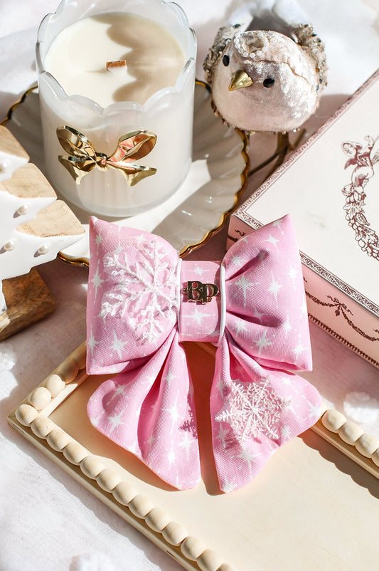 Pink decorative bow with gold accents on a white surface, next to a candle and small bird figurine. Snowflake pink cotton dog bow from premium handmade high quality bows for pets. Bop Pop Pets
