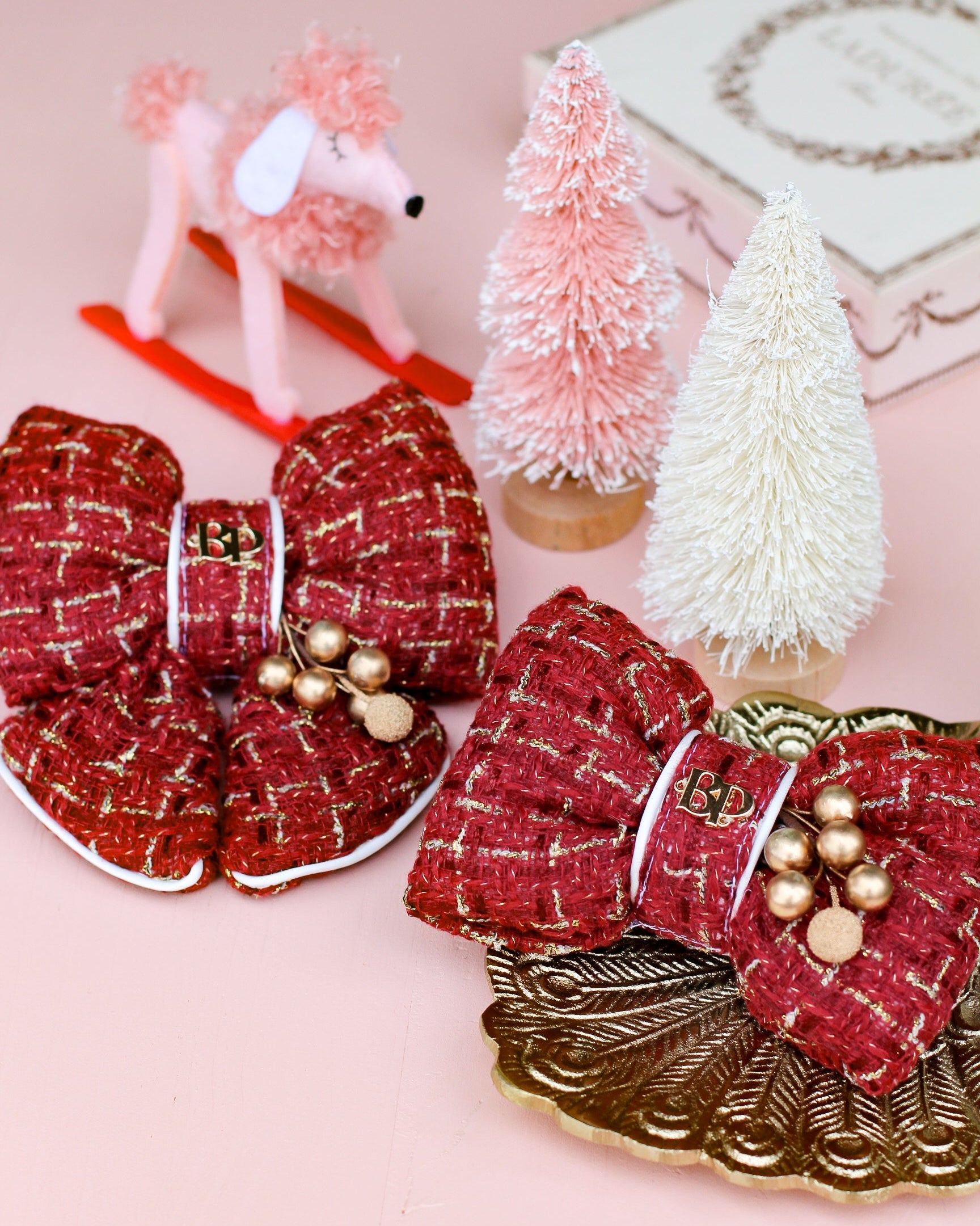 Tweed Burgundy Red decorative sailor bow and bow tie with gold holly accents on a pink table with small Christmas trees and a dog figurine. Holiday picture perfect dog accessory for christmas family portraits. Bop Pop Pets pet accessories