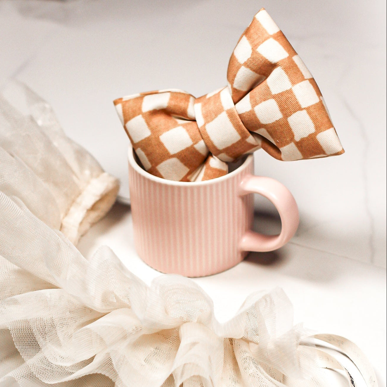 Tan maple checkered bow tie in a pink espresso cup on a marble surface. Bop Pop Pets pet accessory.