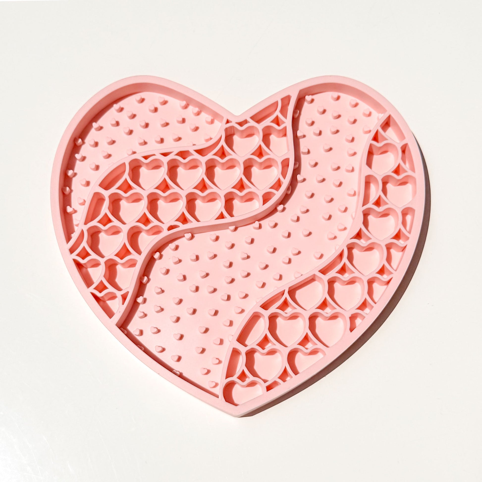 A heart-shaped pink silicone pet lick mat with textured and patterned sections, photographed on a clean white background.