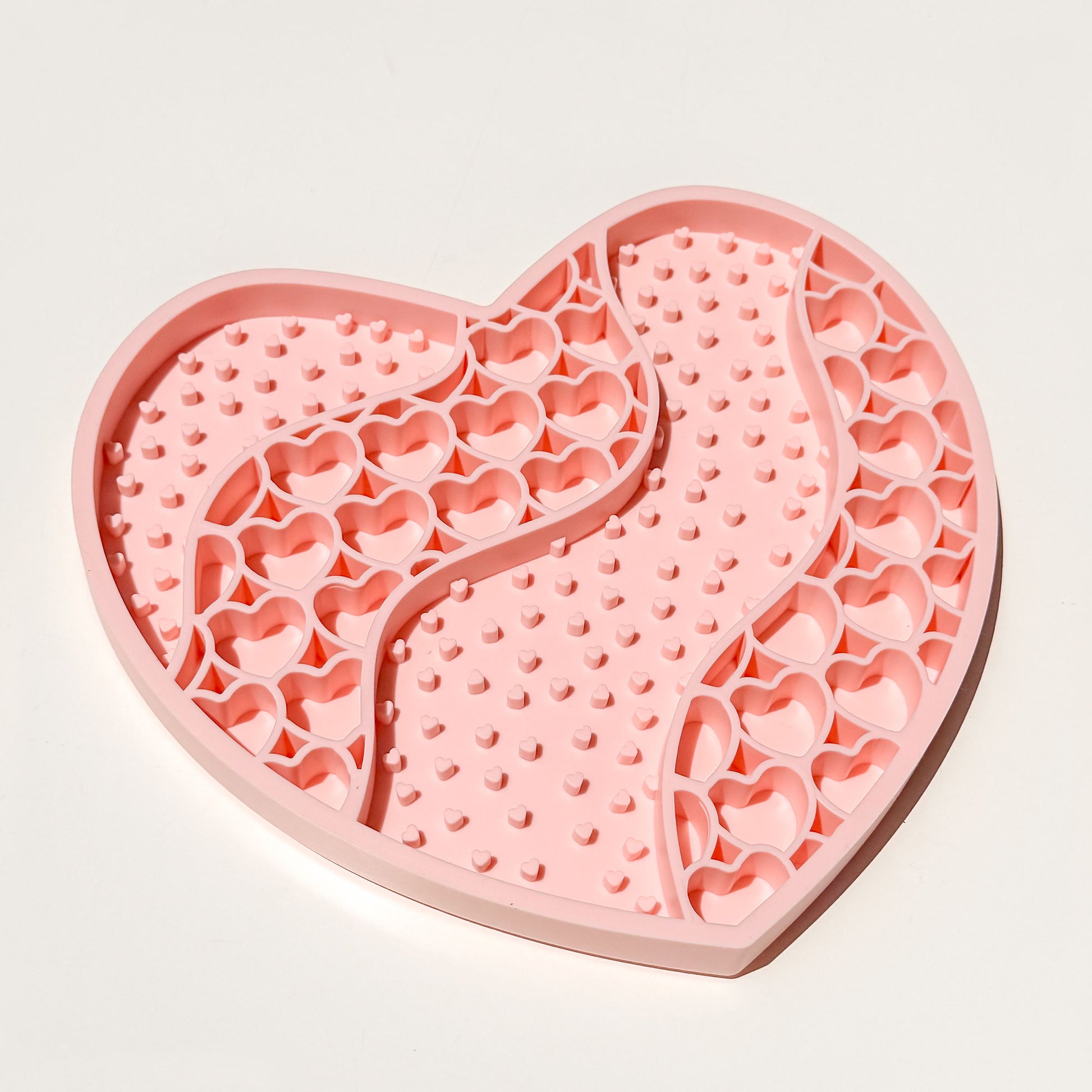 A pink heart-shaped silicone lick mat with a wavy center divider and heart-shaped recesses, photographed on a clean white background.