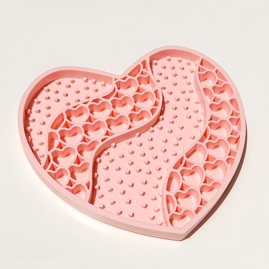A pink heart-shaped silicone lick mat with a wavy center divider and heart-shaped recesses, photographed on a clean white background.