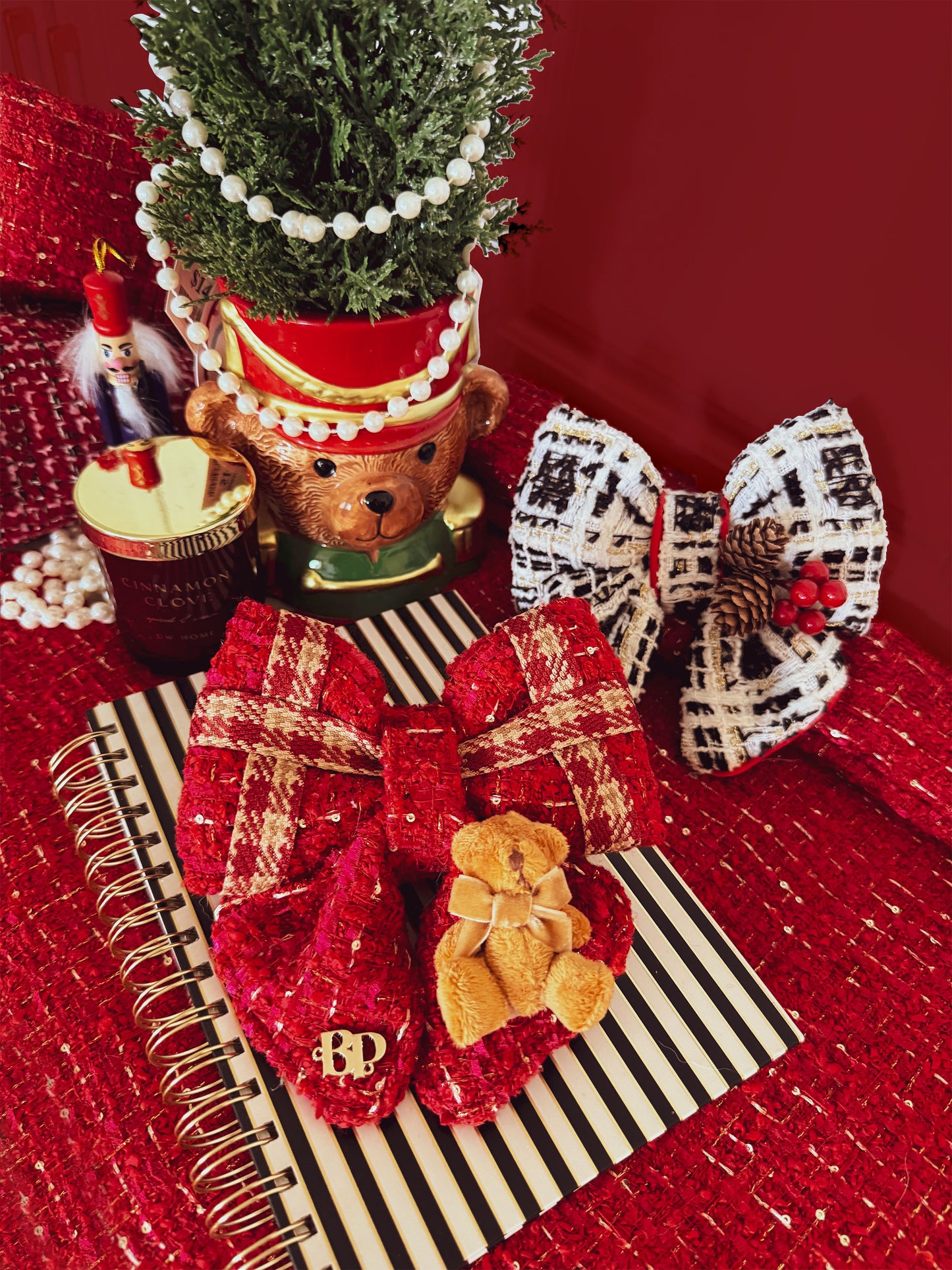 A styled holiday flat-lay featuring a red textured bow with a small teddy bear accent on a black-and-white striped notebook, alongside other festive decorations including a nutcracker, a bear-themed pot planter, and another patterned bow against a deep red background.
