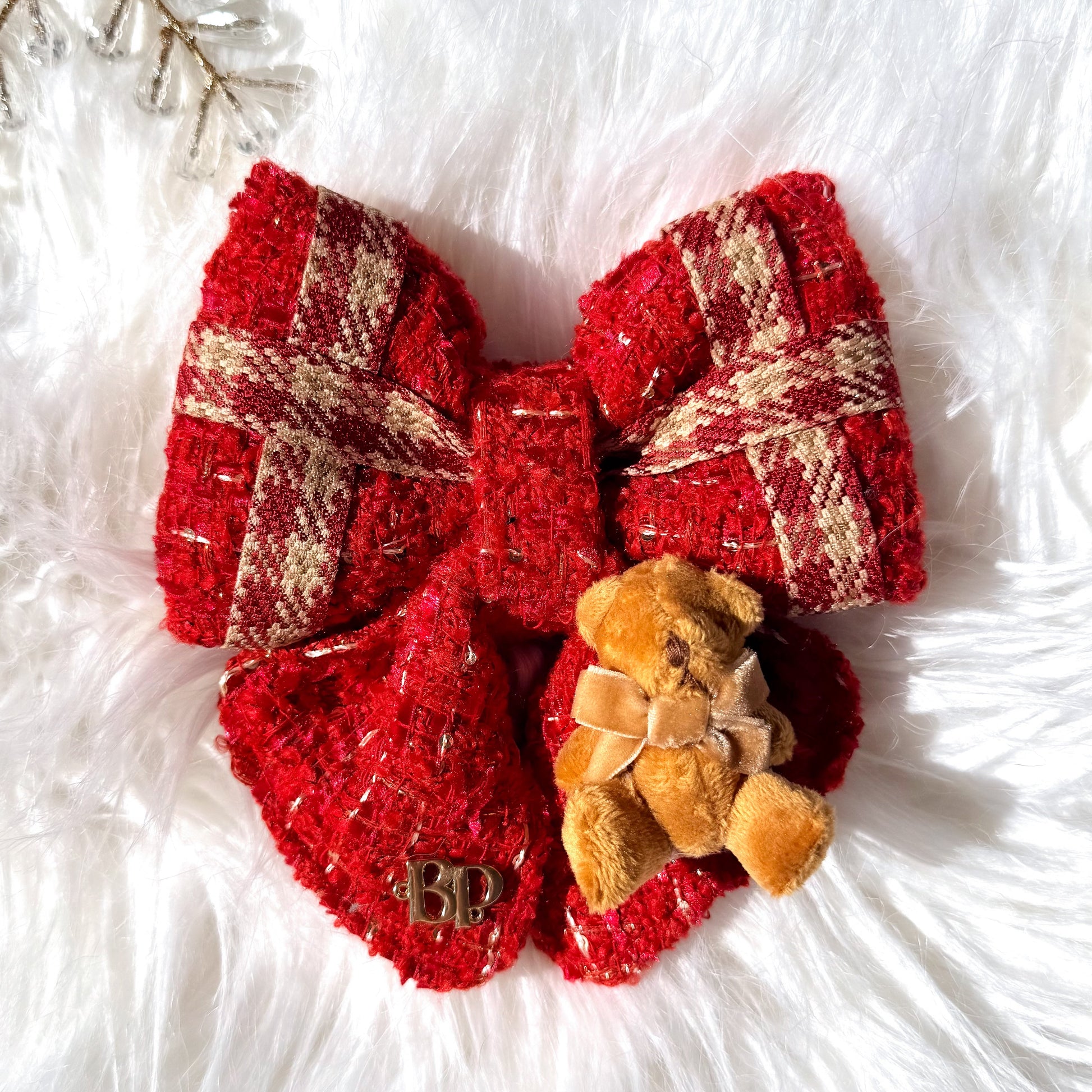 A red and beige plaid textured pet bow with a small brown teddy bear accessory, displayed on a white faux fur background with a decorative gold snowflake.