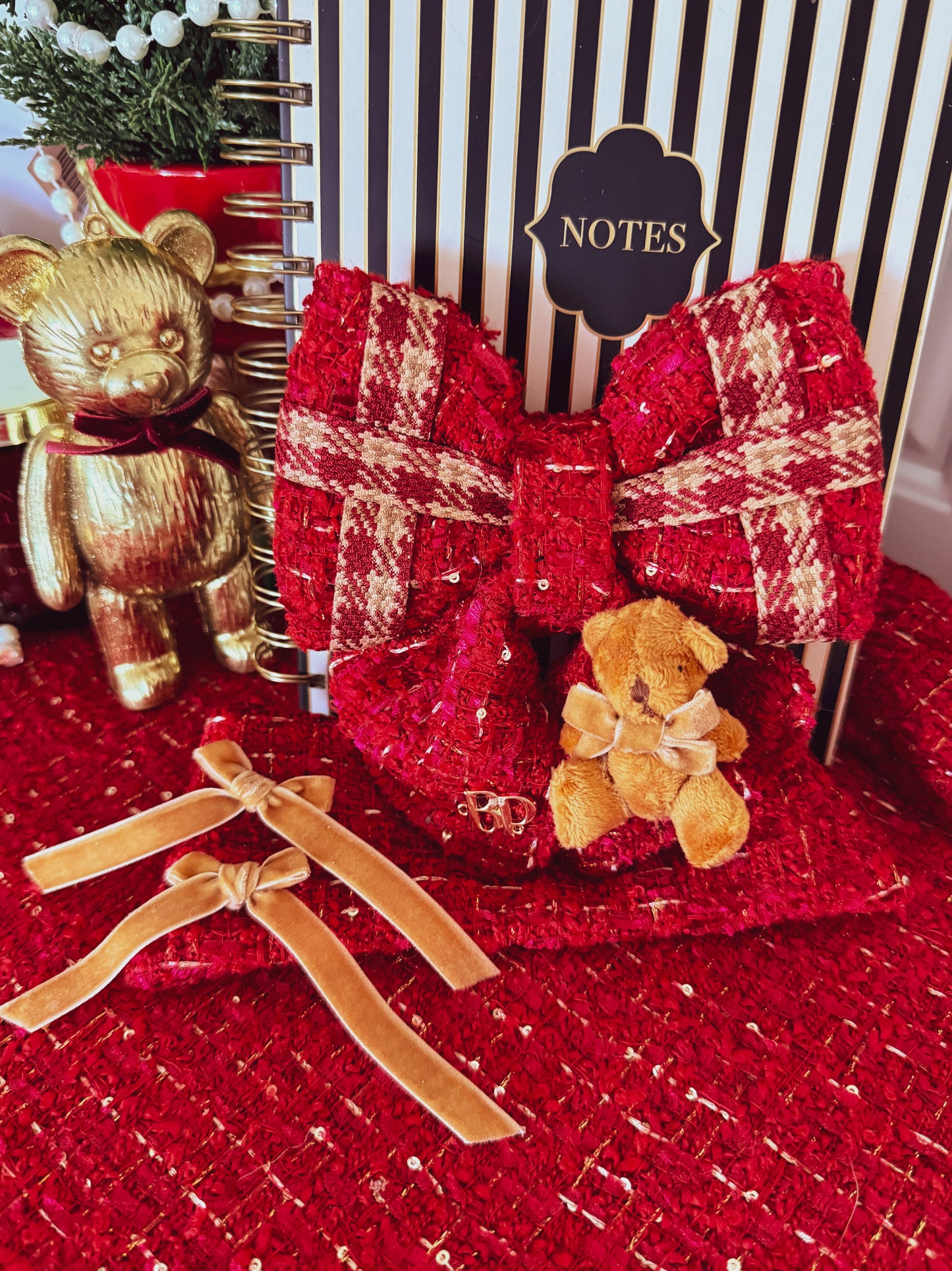 A red tweed notebook with a large red and beige plaid bow, a small teddy bear accent, and two tan ribbon bows on a matching red tweed surface, styled with a gold teddy bear figurine and a striped notebook in the background.