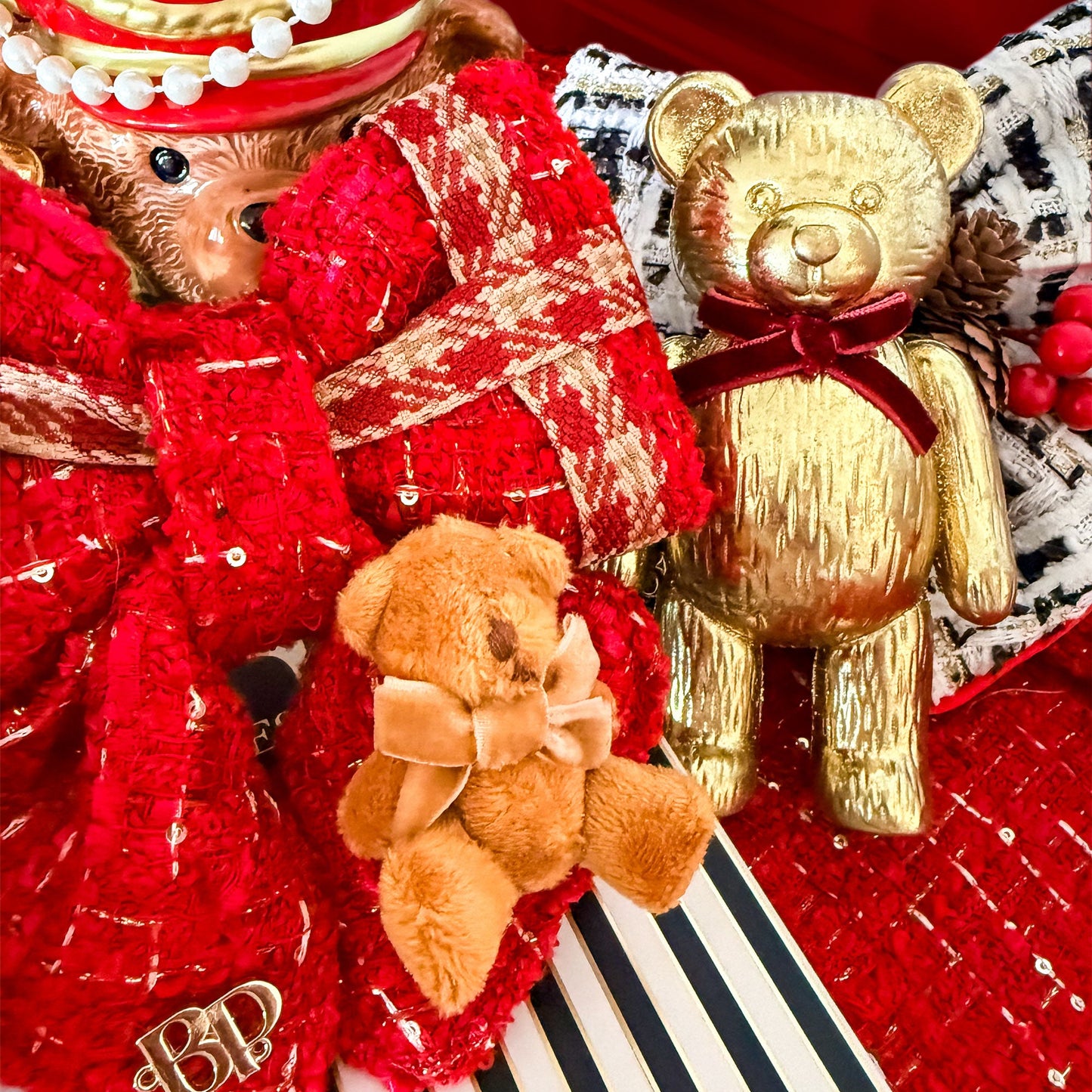A festive holiday-themed product display featuring a plush teddy bear in a red and gold hat, a large red sequined bow, a small brown teddy bear with a beige bow, and a metallic gold teddy bear wearing a dark red ribbon, arranged on a black-and-white striped surface with red textured fabric in the background.
