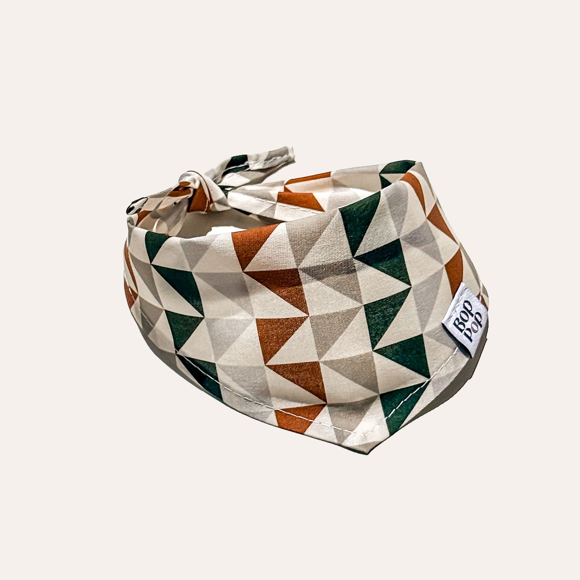 A fabric pet bandana with a geometric triangle pattern in rust orange, dark green, and cream, featuring a small white brand tag that reads 'BOP POP'.