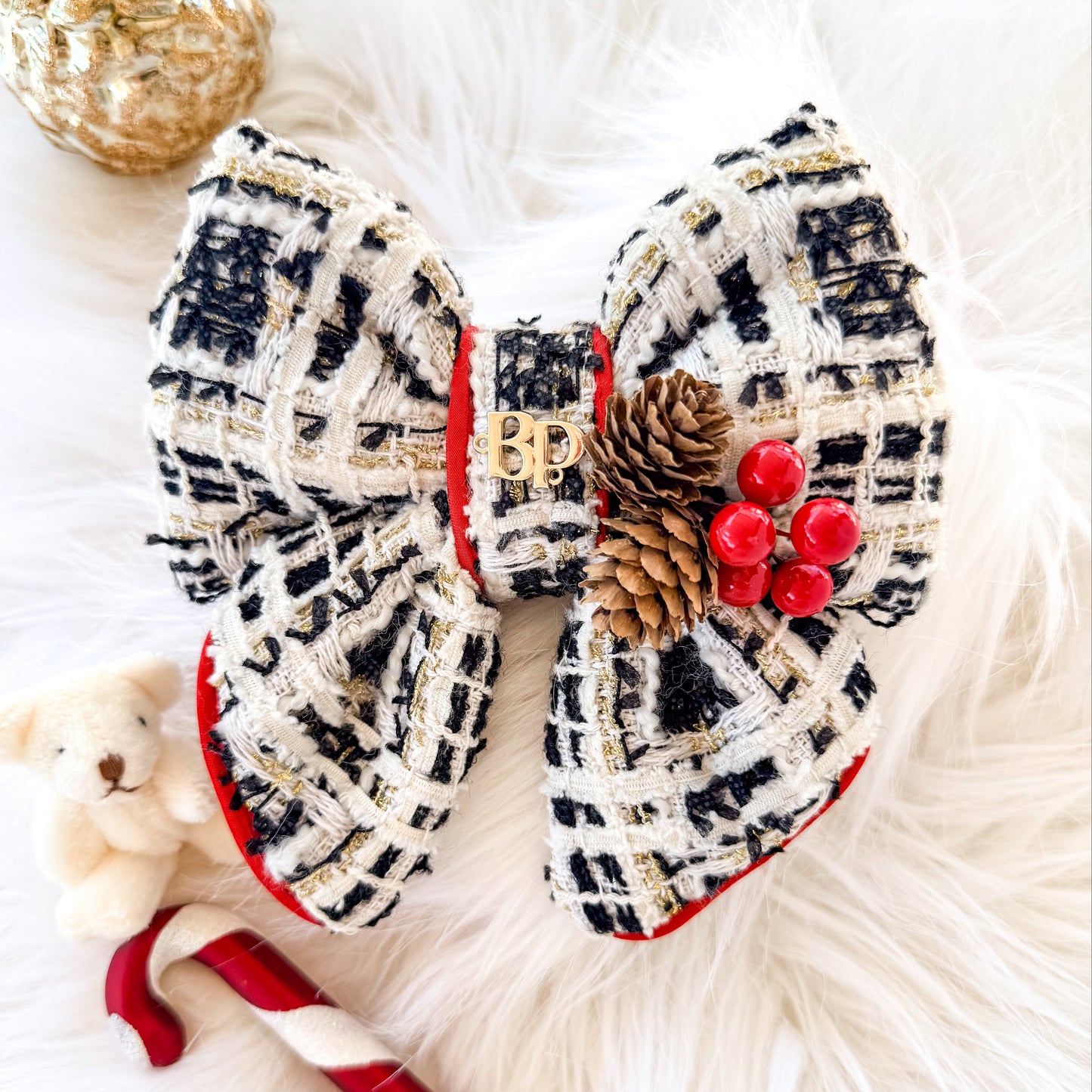 Puffy holiday-themed pet bow tie made of black and beige tweed fabric with red satin trim, decorated with a small pine cone and red holly berryies, styled on a white faux fur background with a teddy bear, candy cane, and gold ornament.