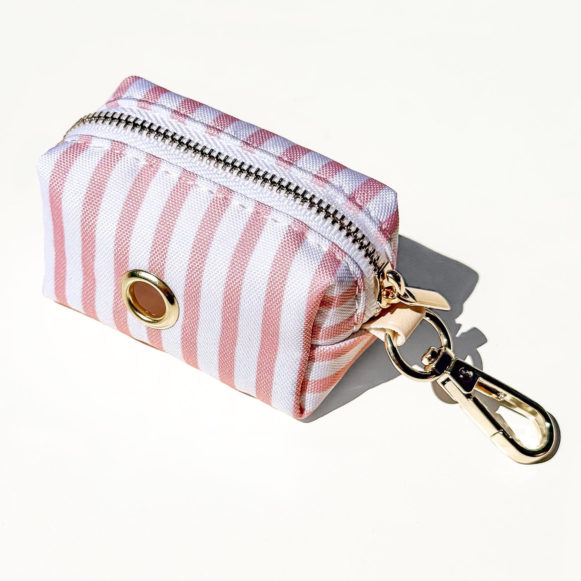 A small zippered pouch with vertical pink and white stripes, featuring a gold-tone metal open grommet and a gold-tone carabiner clip, shown on a clean off-white background.