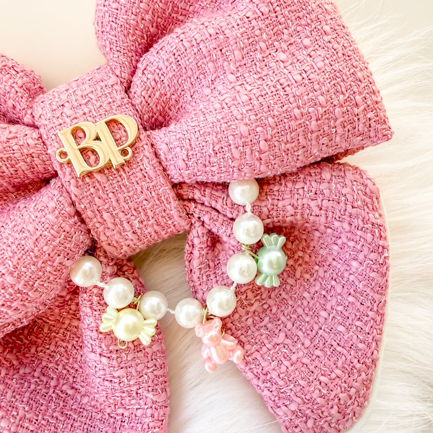 Close up image of pink tweed-like fabric of fancy dog bow tie with tails and  a gold 'BP' brand charm with a dangling pearl and pastel bead chain, displayed on a white faux fur surface.