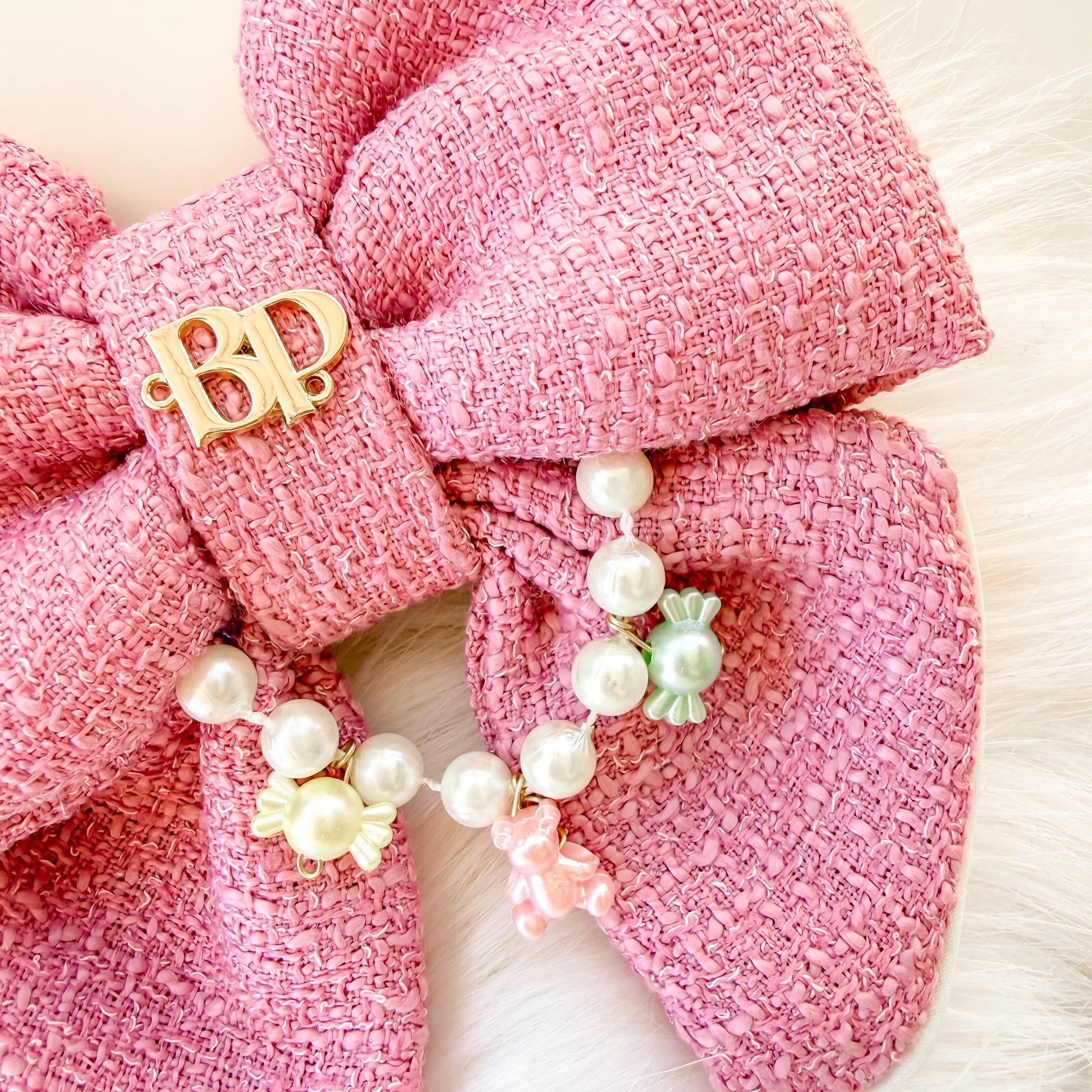 Close up image of pink tweed-like fabric of fancy dog bow tie with tails and  a gold 'BP' brand charm with a dangling pearl and pastel bead chain, displayed on a white faux fur surface.