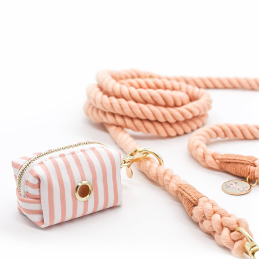 A striped pink and white poop bag holder with a gold-colored snap-on carabiner and zipper closure attached to a pink salmon colored dog rope leash with gold hardware and circle tag with pink dog branding.