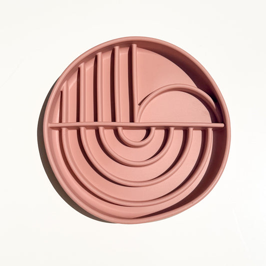 A round, dusty pink silicone pet slow feeder bowl with concentric wavy ridges and vertical slits, shown from a top-down perspective on a clean white background.