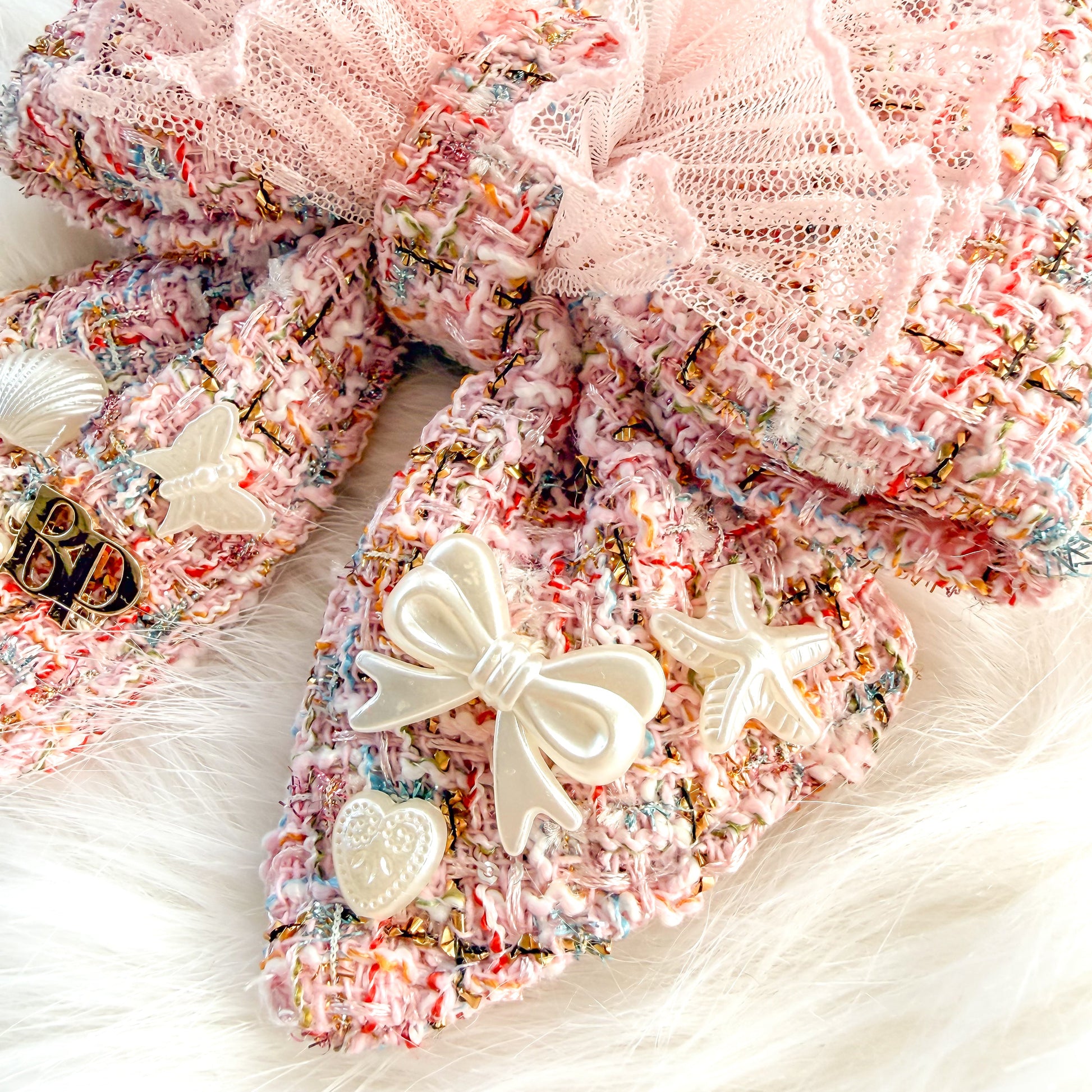 A large decorative pet bow tie with tails made from pink tweed fabric with a sheer light pink ruffled overlay, styled on a white faux-fur surface with holiday pearl-white ornaments and small embellishments.