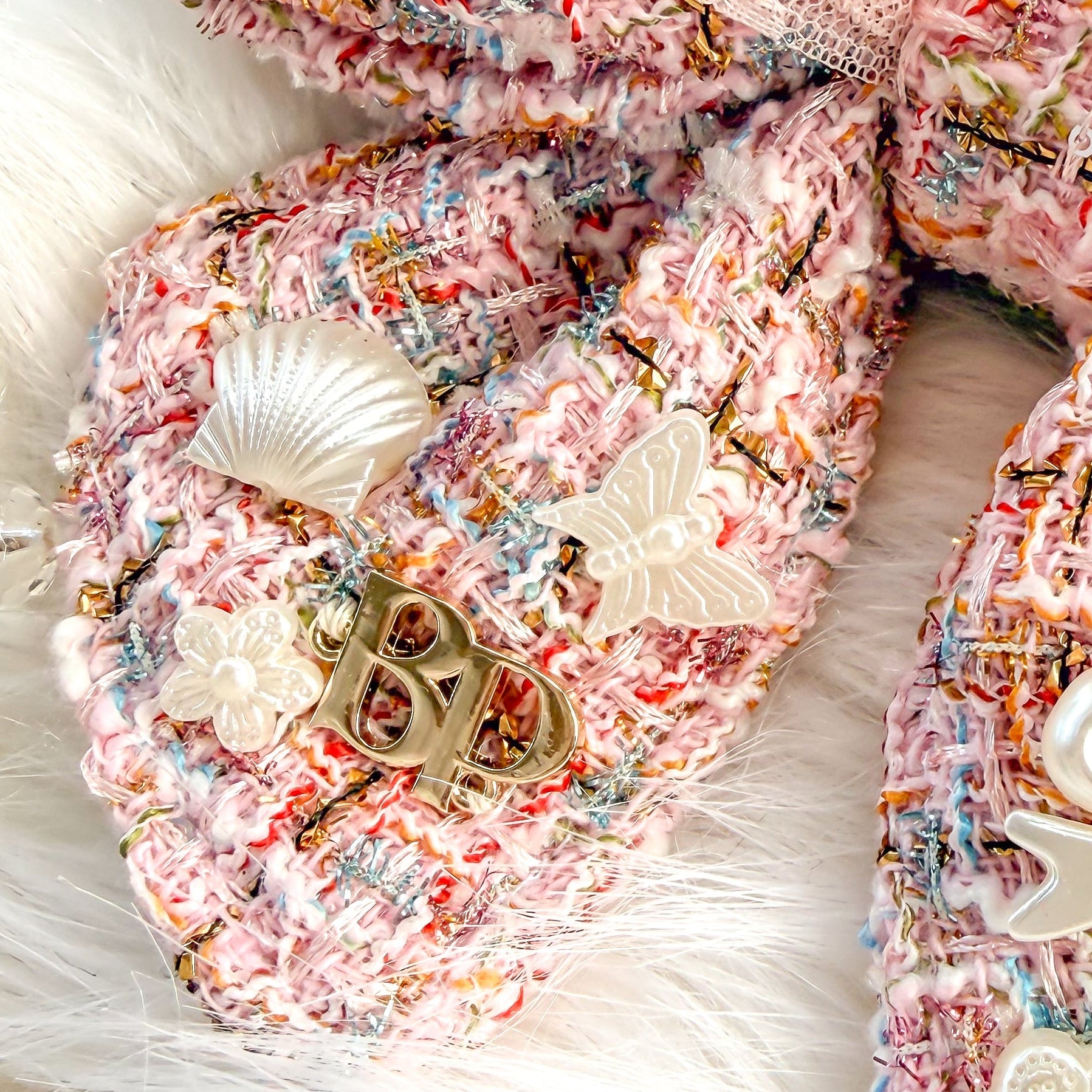 A large decorative pet bow tie with tails made from pink tweed fabric with a sheer light pink ruffled overlay, styled on a white faux-fur surface with holiday pearl-white ornaments and small embellishments.