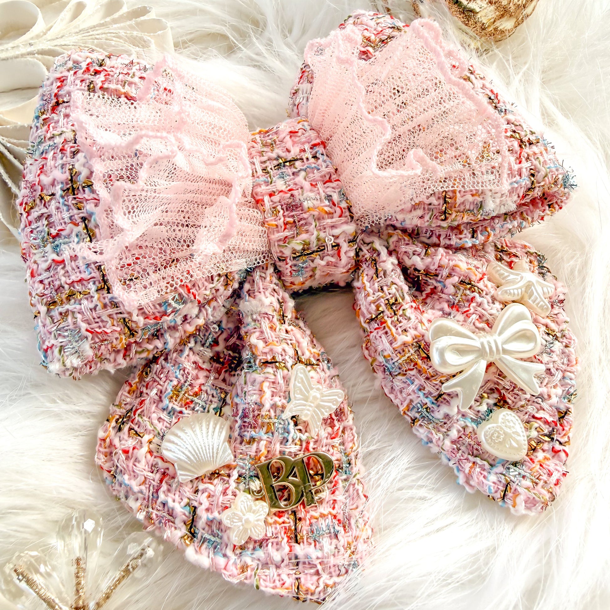 A large decorative pet bow tie with tails made from pink tweed fabric with a sheer light pink ruffled overlay, styled on a white faux-fur surface with holiday pearl-white ornaments and small embellishments.