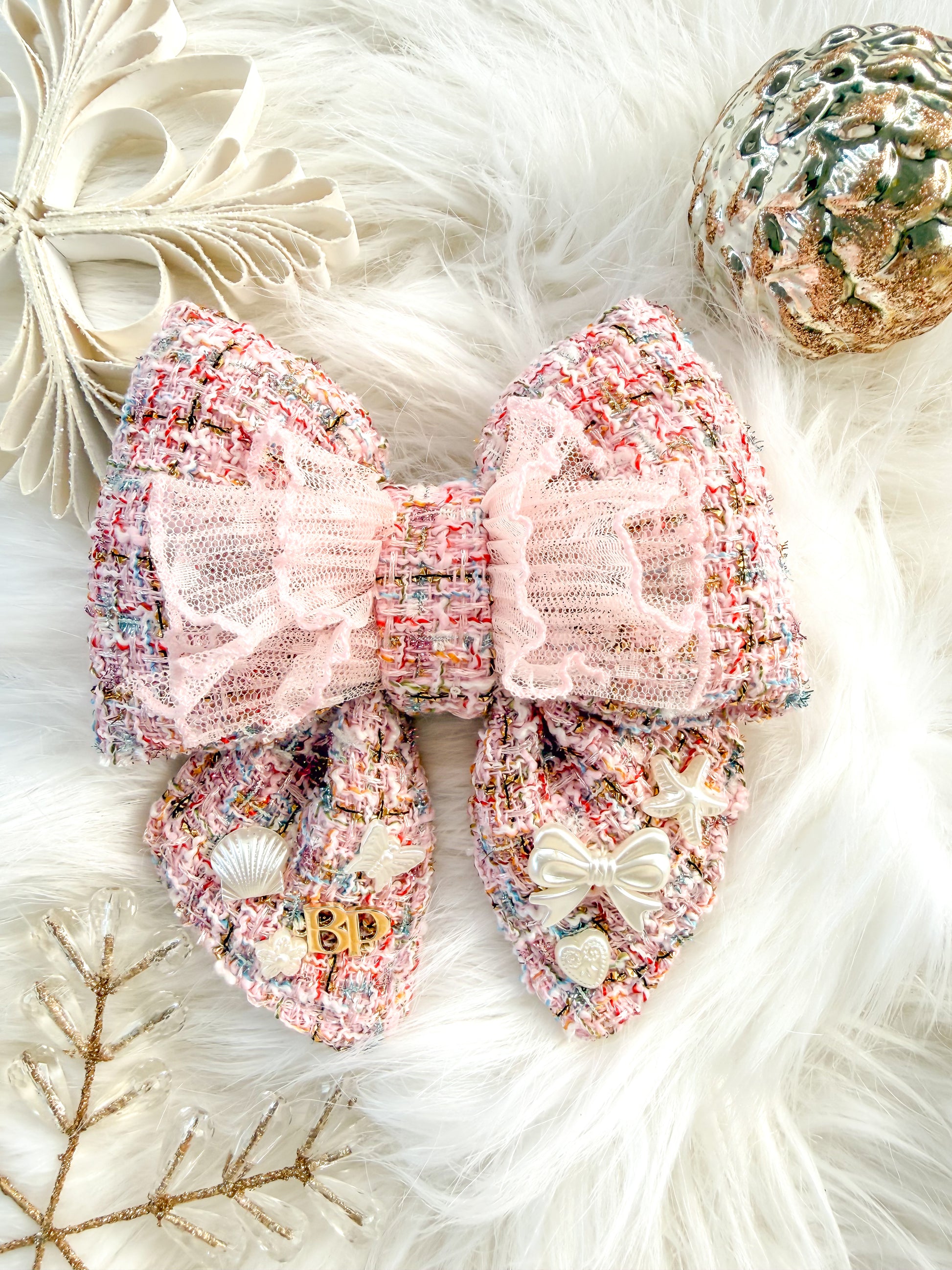 A large decorative pet bow tie with tails made from pink tweed fabric with a sheer light pink ruffled overlay, styled on a white faux-fur surface with holiday pearl-white ornaments and small embellishments.