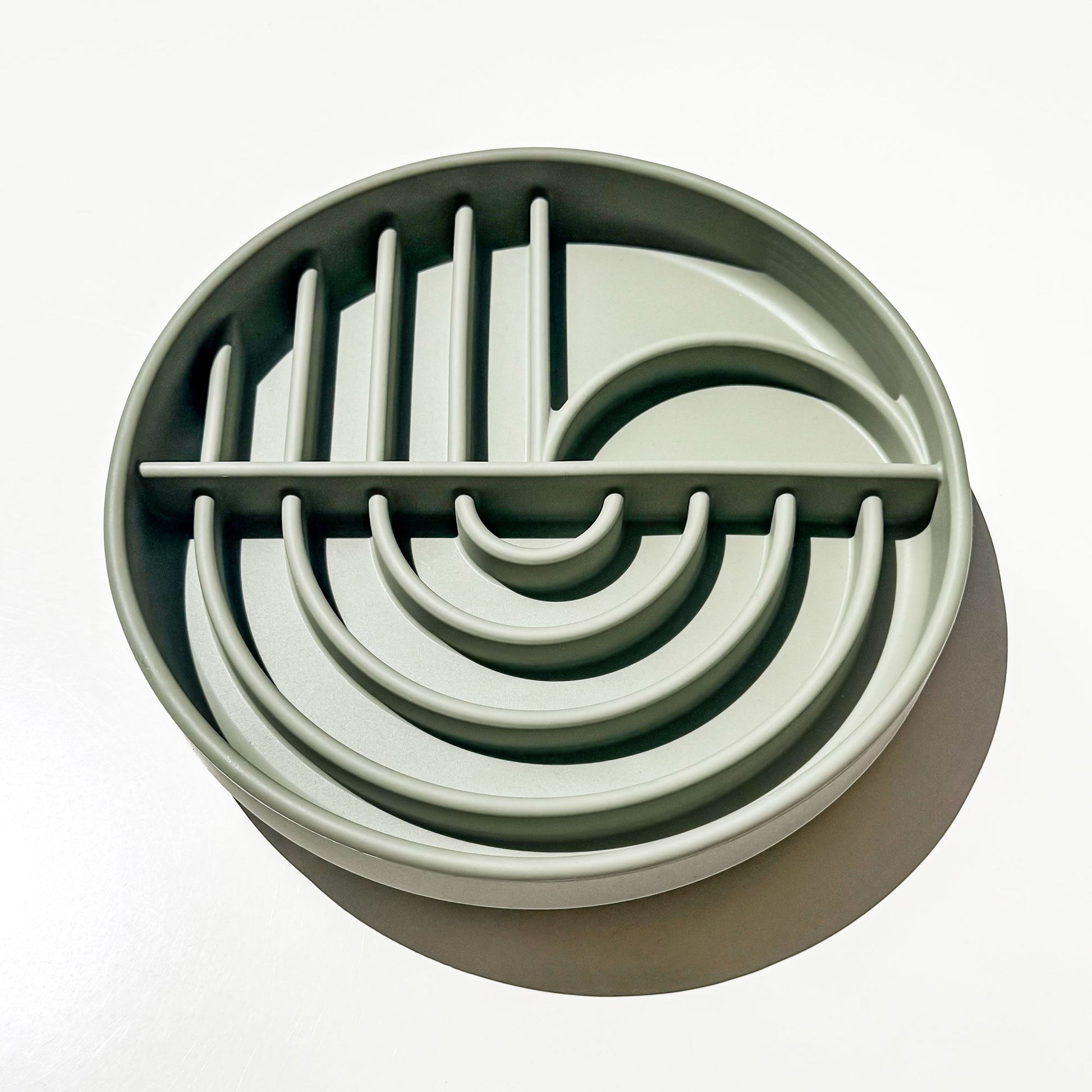 A round, light sage green silicone pet slow feeding deep dish bowl with geometric arch division pathways and divider slots for enrichment feeding, shown on a clean white background with a soft shadow.