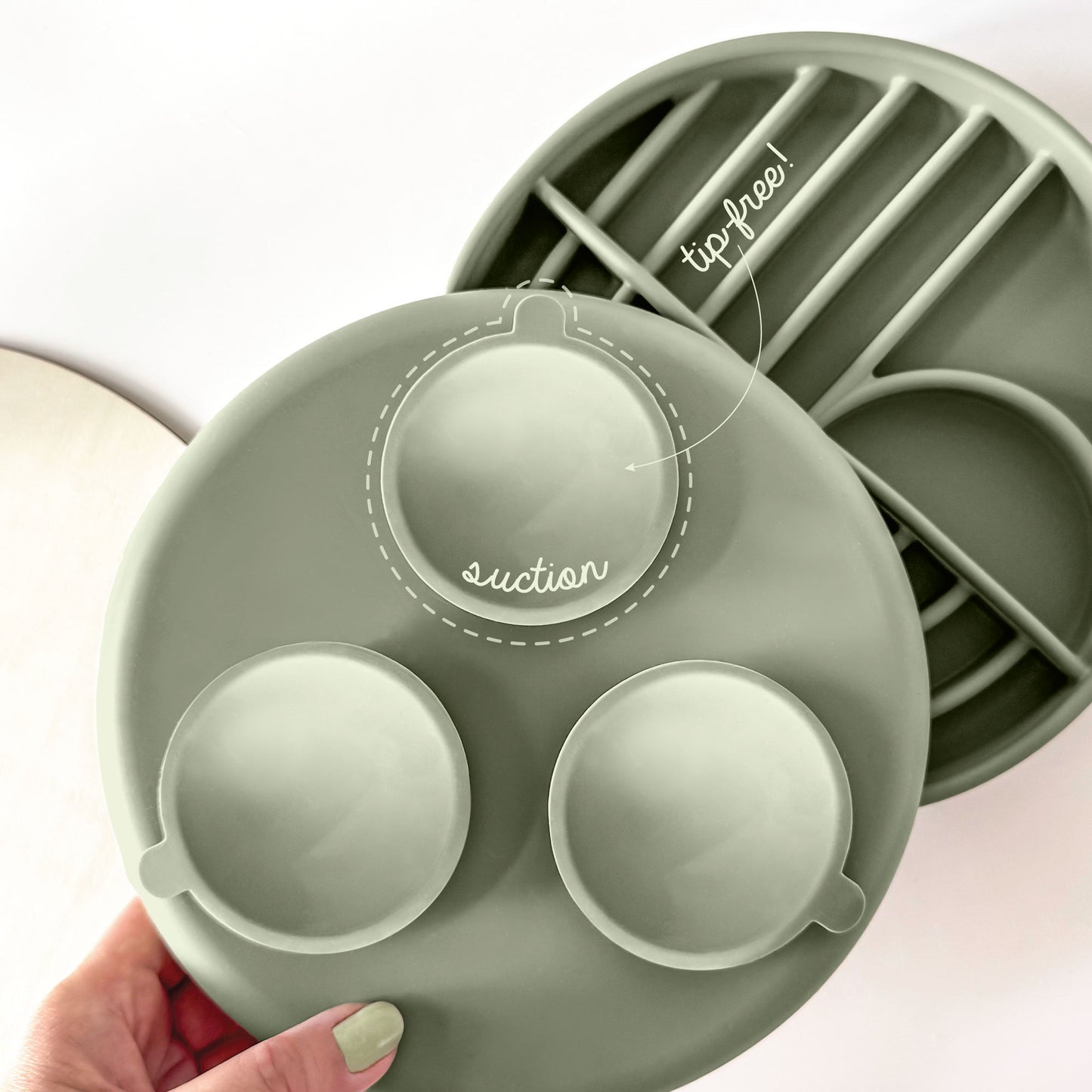 A sage green silicone baby feeding plate with suction cups and a divided tray, held by a hand. A second view of the plate showing its divided compartments is in the background, with text overlays indicating 'tip-free!' and 'suction'.