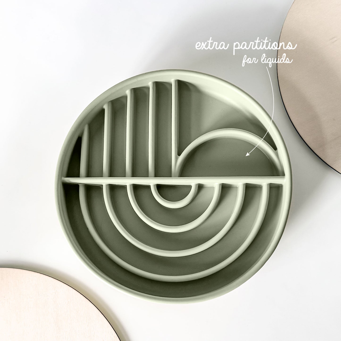 A round, light sage green silicone pet slow feeding shallow bowl with geometric compartments, shown on a white background with decorative wooden plates and text overlay 'extra partitions for liquids'.
