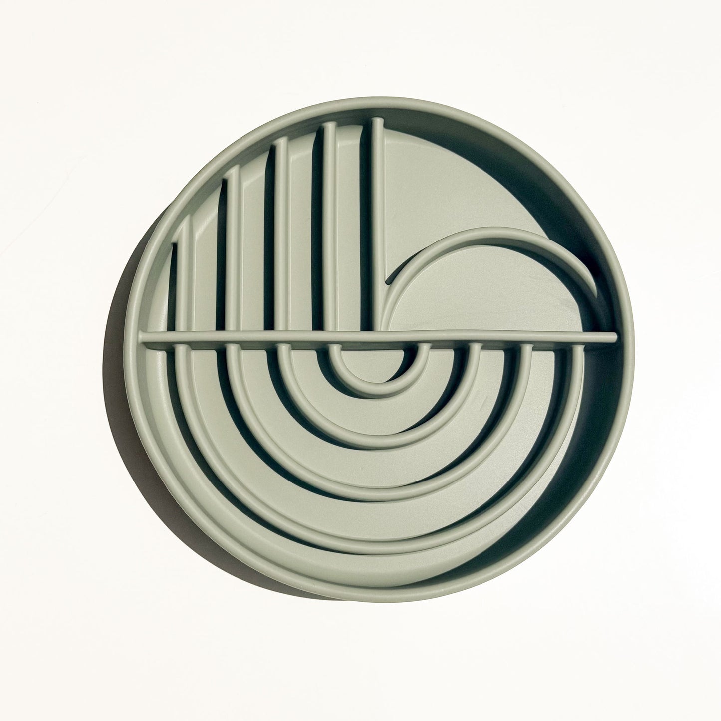 A round, light sage green silicone pet slow feeder shallow bowl with a boho style maze pattern of concentric arches, vertical bars, and a curved loop, shown from a top-down perspective on a white background.