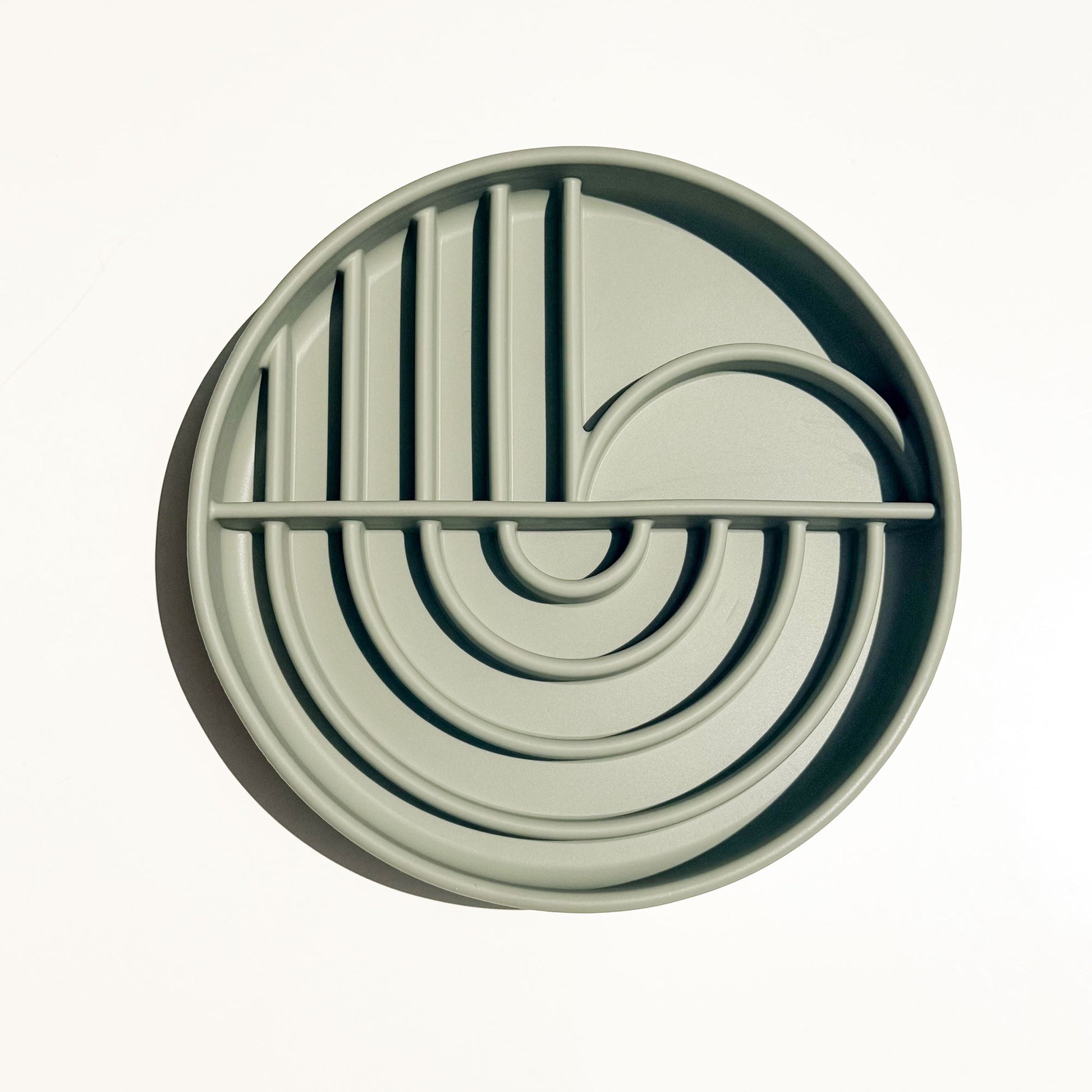 A round, light sage green silicone pet slow feeder shallow bowl with a boho style maze pattern of concentric arches, vertical bars, and a curved loop, shown from a top-down perspective on a white background.