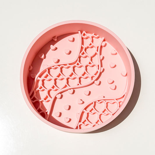 A round pink blush colored silicone pet slow feeder bowl with a wavy pattern of hearts small and large, photographed from directly above on a white background.