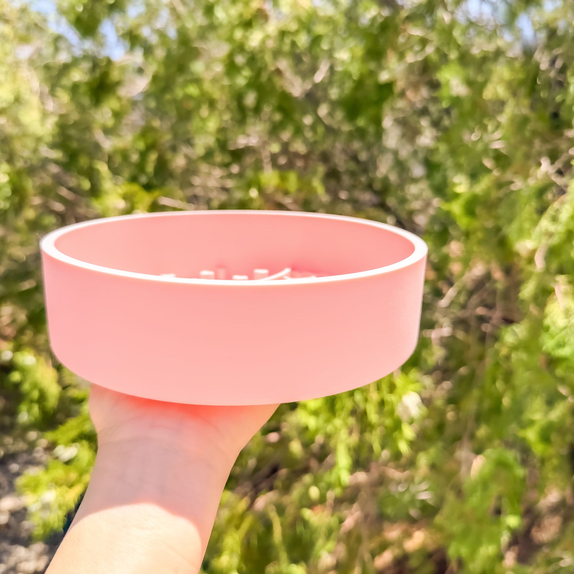 A light pink silicone pet slow feeder bowl held by a hand against a blurred background of green foliage.