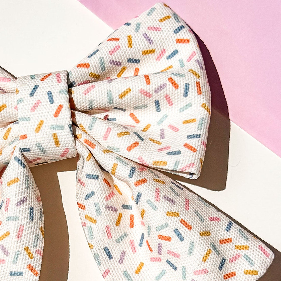 Close up detail of pet bow with a white base and a colorful sprinkle pattern, featuring a small gold 'BP' monogram. It is styled on a white surface with a pink background corner and a decorative gift box.