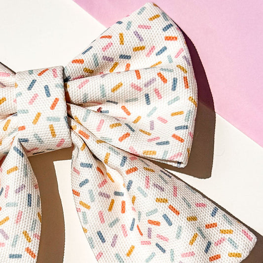 Close up detail of pet bow with a white base and a colorful sprinkle pattern, featuring a small gold 'BP' monogram. It is styled on a white surface with a pink background corner and a decorative gift box.
