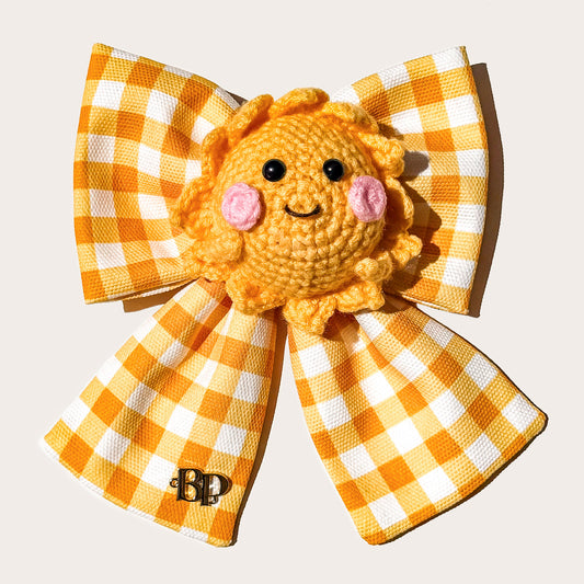 A yellow and white gingham pet bow with tails featuring a crocheted sun character in the center with pink cheeks and black eyes with a BP branded metal logo on left bow tail photographed on a white background.