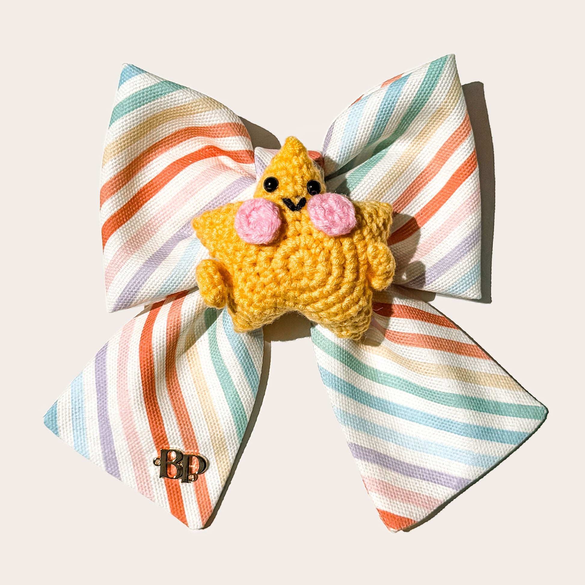 A colorful pastel striped cotton canvas fabric pet bow with a yellow-orange crocheted star character accessory in the center, photographed on a plain white background.