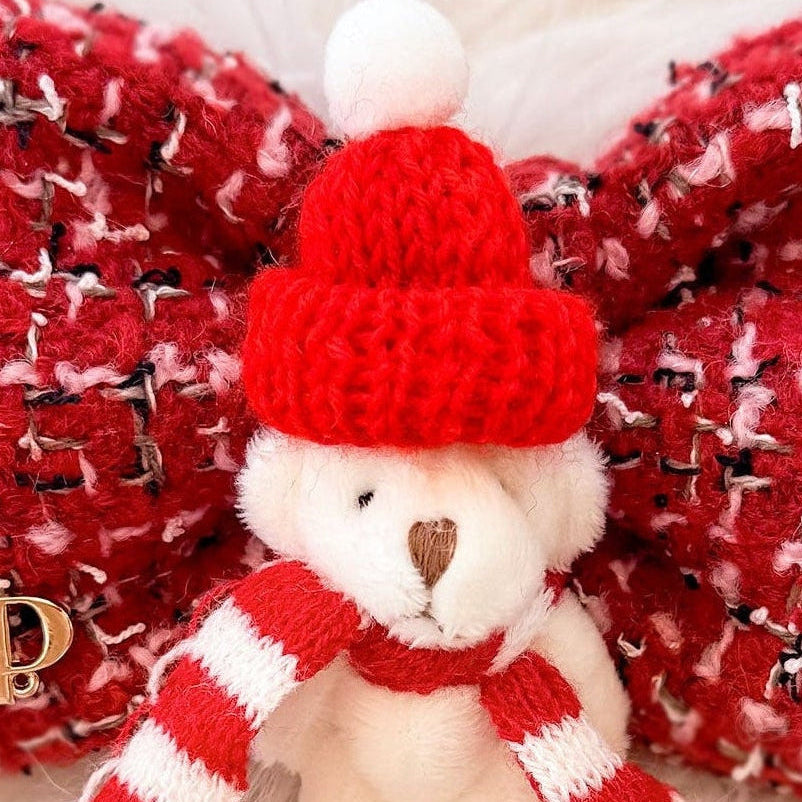 A festive red and white tweed pet bow tie featuring a small white mini plush teddy bear wearing a red knit hat and striped scarf, displayed on a white faux fur surface with holiday greenery.