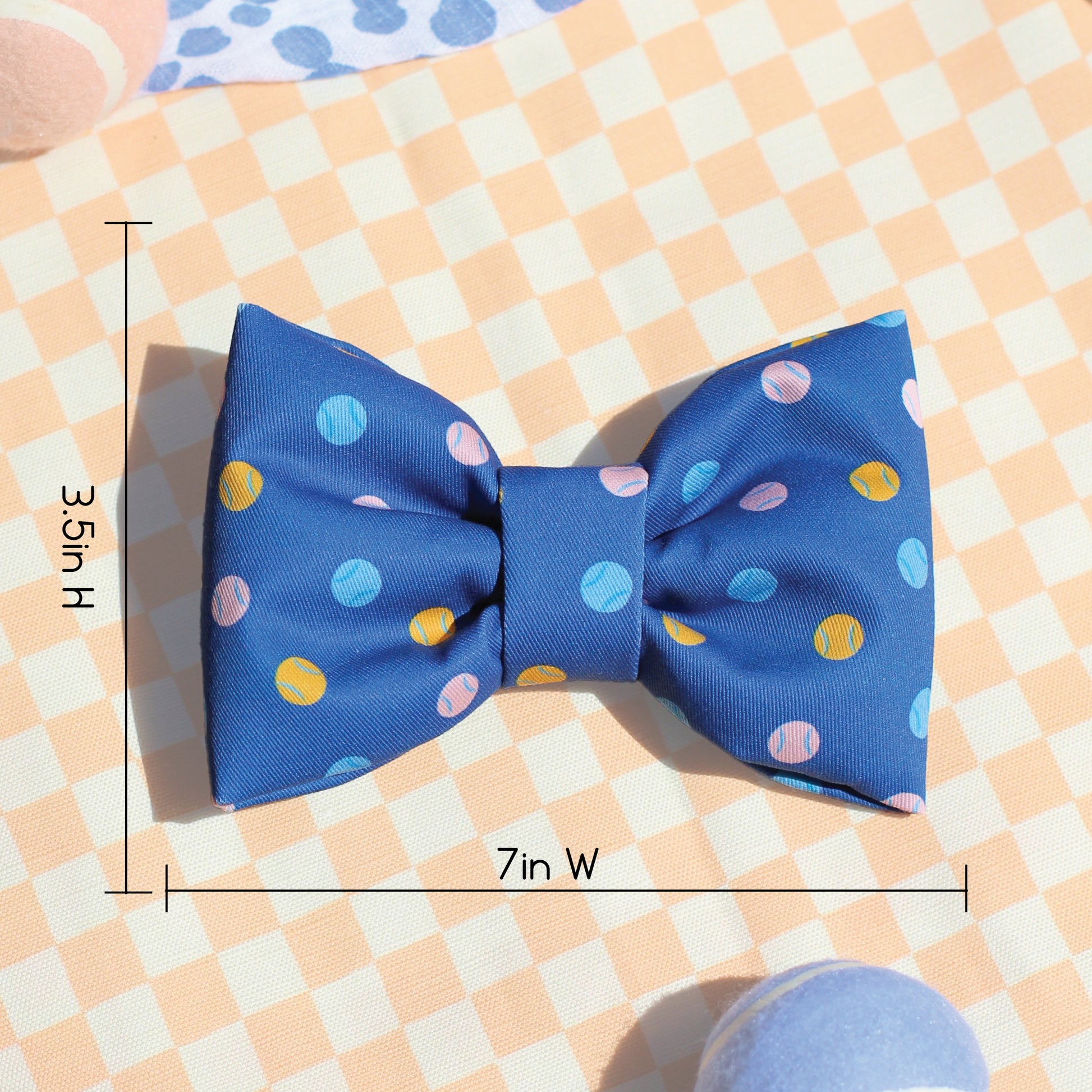 Description of 3.5in H by 7in Wide dimensions for a blue pet bow tie with colorful tennis balls with on a peach checkered fabric background for Bop Pop Pets. Made 100% cotton, puffy bow ties for fashion, trendy dogs and cats 