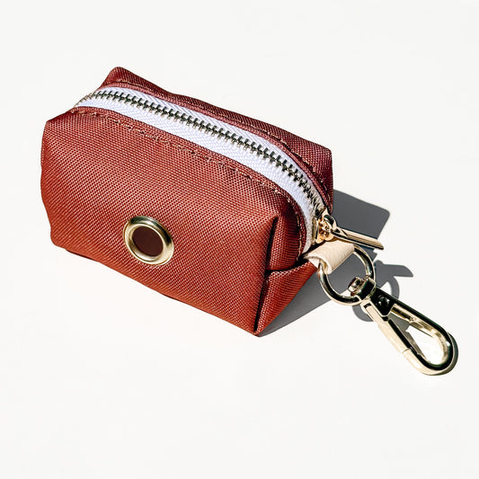 A small reddish-brown canvas zippered pouch with a gold-tone metal grommet and a silver-and-gold carabiner clip, shown on a clean white background.