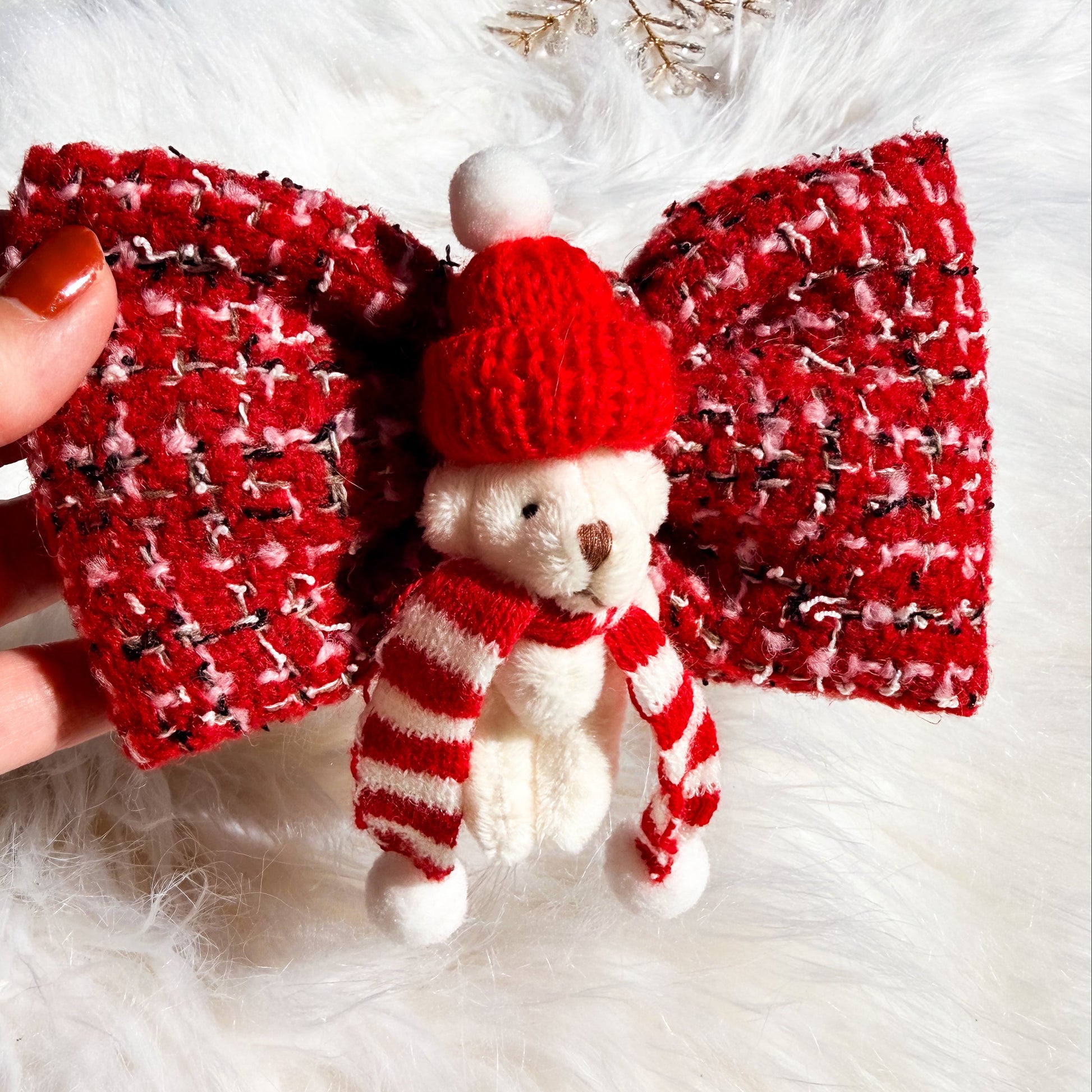 A red and white tweed dog bow accessory with a small knitted teddy bear wearing a red Santa hat and striped scarf, held by a hand over a white faux fur background.