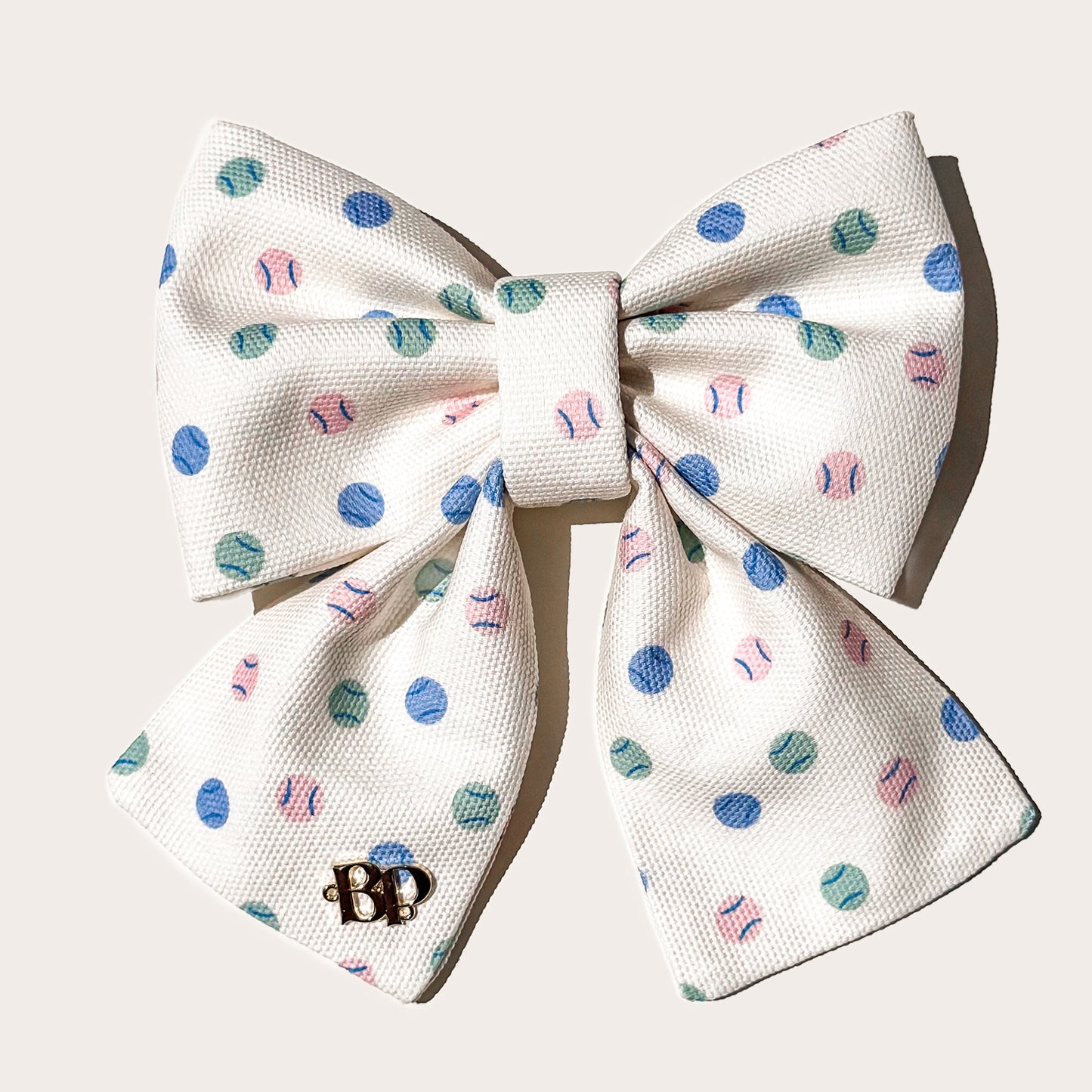 A cotton canvas textured fabric pet sailor bow in cream with a pattern of blue, teal, and pink tennis balls, featuring a small gold 'BP' monogram brand logo on left bow tails on white background.