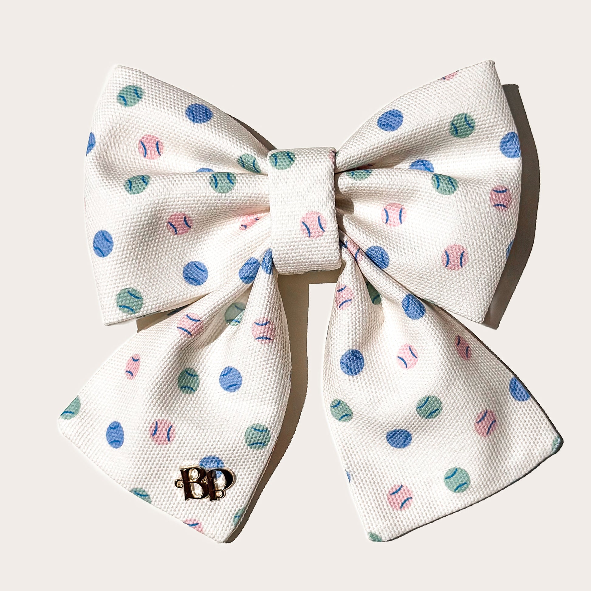 A cotton canvas textured fabric pet sailor bow in cream with a pattern of blue, teal, and pink tennis balls, featuring a small gold 'BP' monogram brand logo on left bow tails on white background.