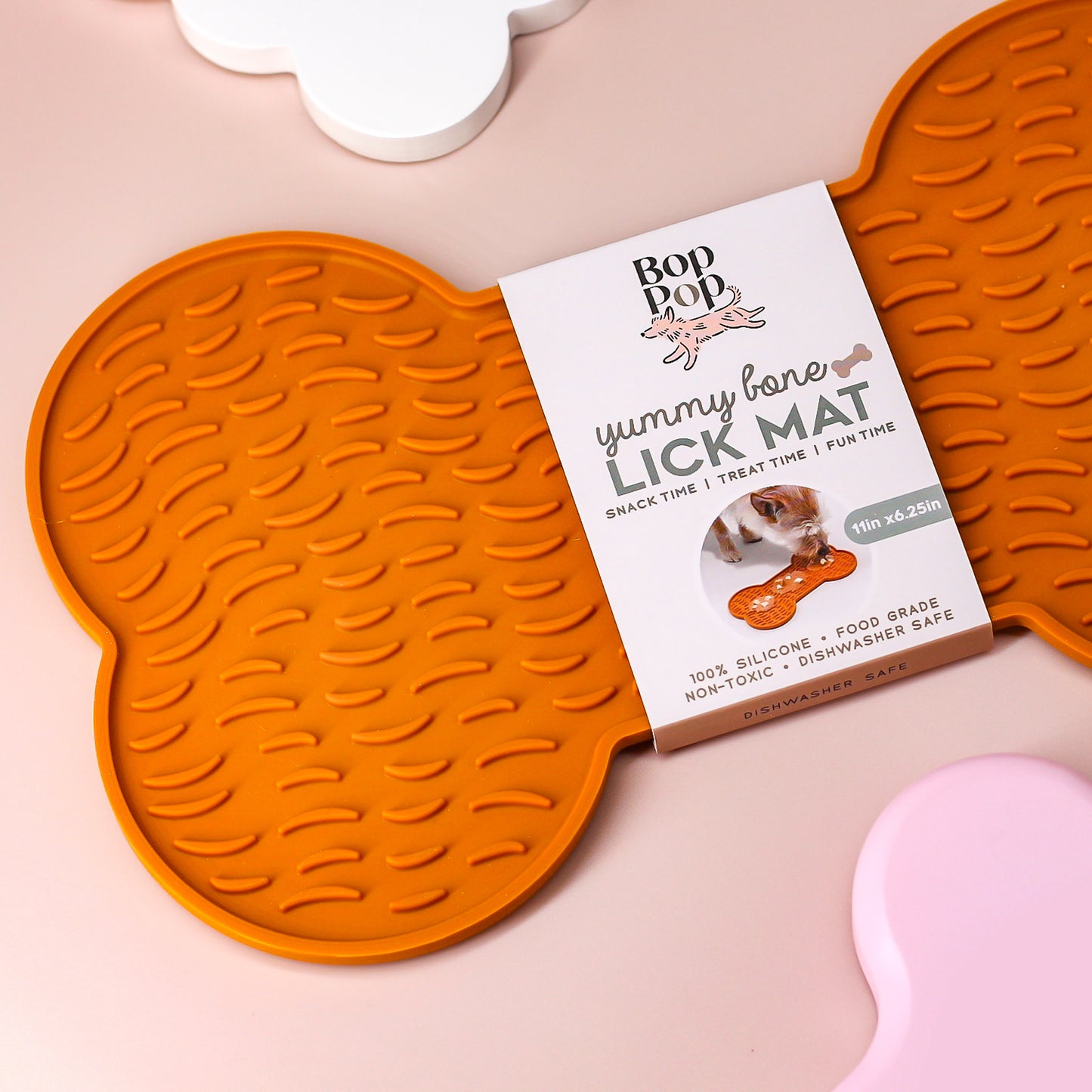 Cropped on right, view of a bone-shaped Lick Mat on a tan colored background for Dogs or Cats made of silicone material in burnt orange with packaging displayed with Bop Pop brand logo with peach dog icon.