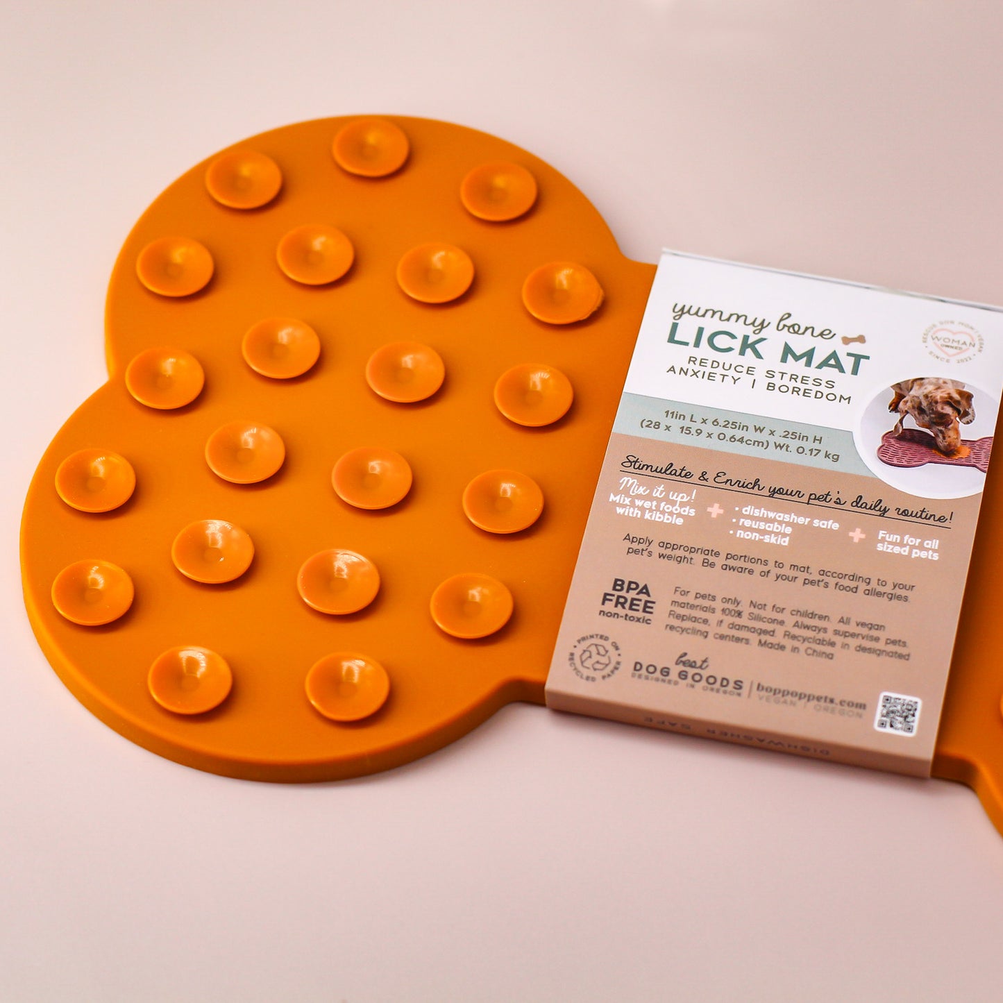 Cropped on right, back view of a bone-shaped Lick Mat on a tan colored background  made of silicone material in burnt orange with packaging displayed with Bop Pop brand logo with peach dog icon. Display shows details of small circle suctions on back of lick mat and size details, BPA free and color photo of Dog licking a mat usage.